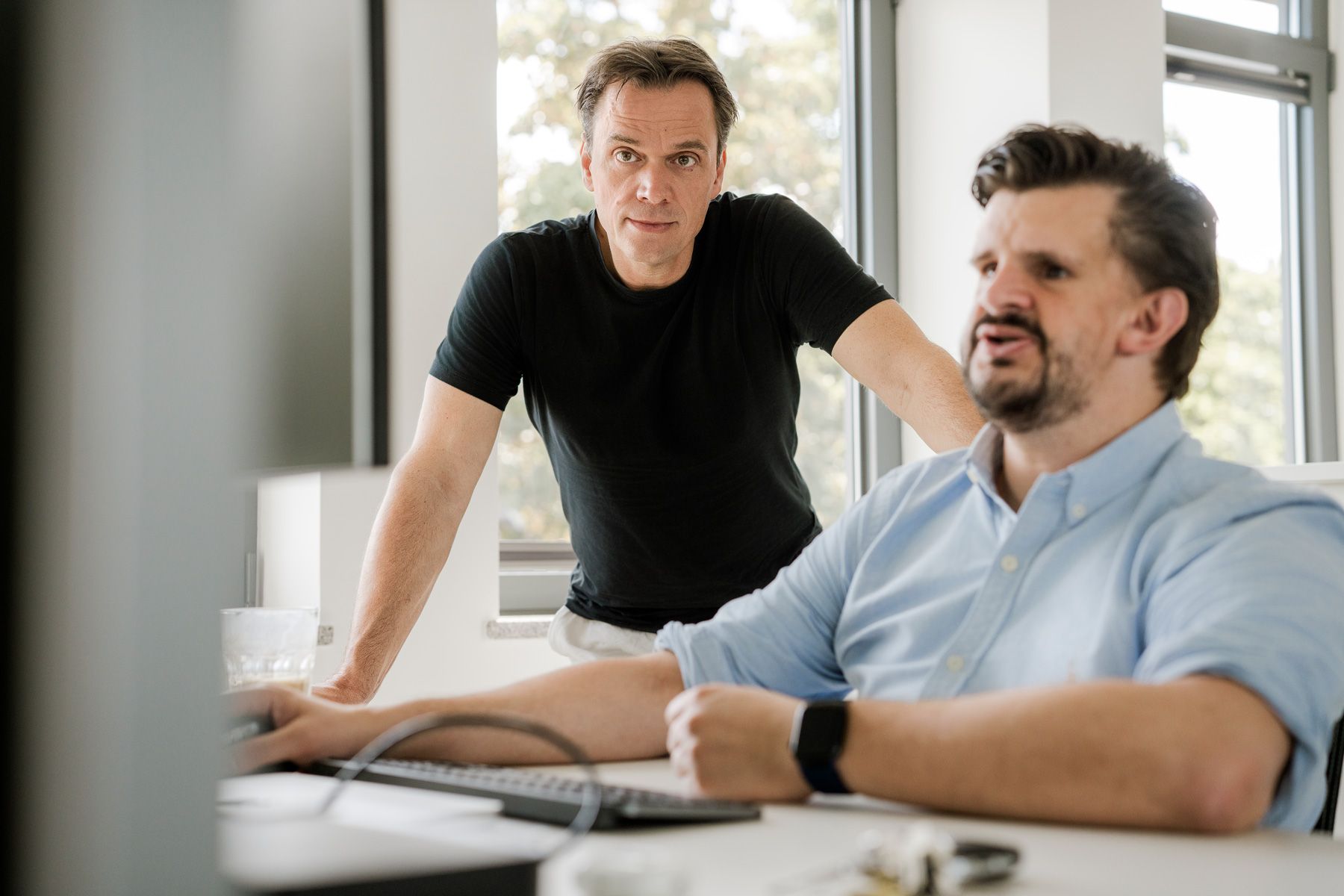 Two people are standing and sitting at a desk, discussing what's on the screen in an office.
