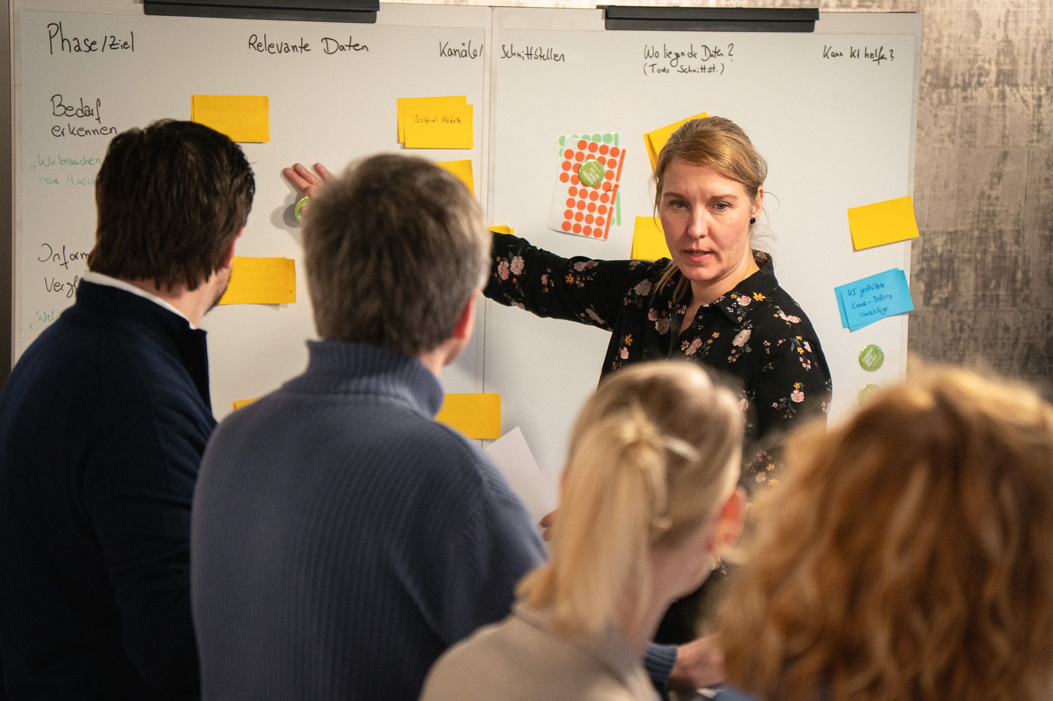 Several people are standing in front of a whiteboard covered with Post-it notes; one person is explaining the content and pointing at the board.